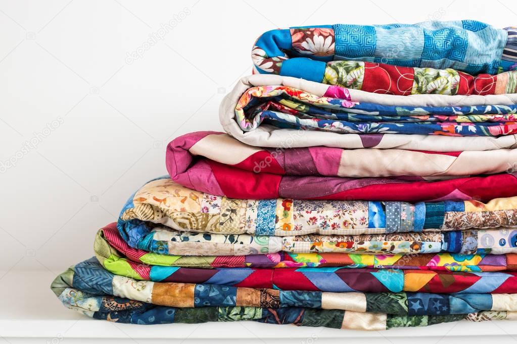 sewing, patchwork and fashion concept - beautiful colorful quilts were neatly folded and stored in several rows in height for storage, sale of finished textile stitched products on white background.