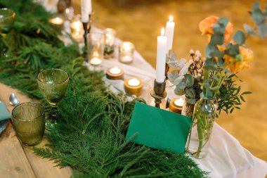 Kış, tasarım, atmosfer konsepti. Masanın dekorasyonu iki aşığın nişanını kutlamak için. Kozalaklı taze zümrüt dallar, mumlar, şeffaf şişelerdeki desenler.