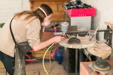 Plastik sanatlar, el yapımı konsept. Gündelik kazak, kot pantolon, iş önlüğü ve solunum cihazı giymiş genç sarışın kız stüdyoda çömlekçilikle çalışıyor.