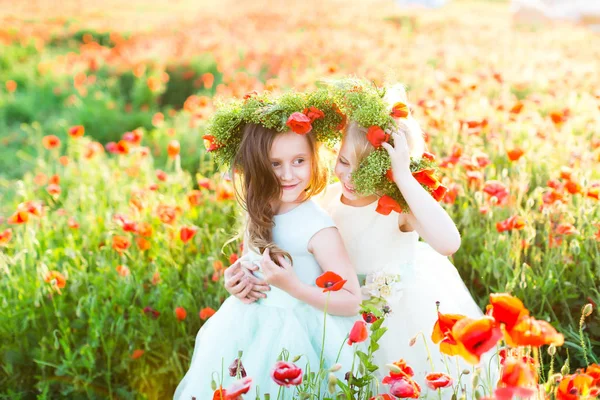 Modelo niña, boda, amapolas, concepto de moda de verano - abrazos de dos niñas en vestidos de lujo brillantes en un campo soleado de amapolas en flor, las dos chicas riendo en coronas de flores de verano — Foto de Stock