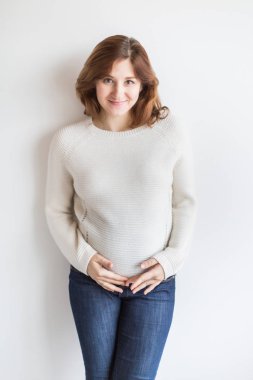 Aşk, sağlık, beklenti konsepti. Gülümseyen bir kadın var. Şık ama sade kıyafetler giyiyor. Hamileymiş ve karnına nazikçe sarılıyormuş.