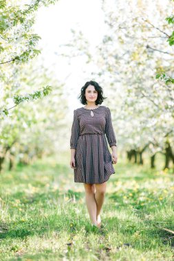 Bahar, doğa, yaşam tarzı konsepti. Kısa siyah saçlı ve açık tenli muhteşem bir kadın var. Çiçek açmış kiraz ağaçlarıyla dolu bir bahçede yürüyor.