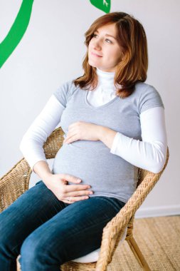 moda, dinlenme, ebeveynlik kavramı. Konforlu ve şık hasır koltukta çok güzel bir anne bekliyor, beyaz yaka kazak giyiyor, gri gömlek ve kot pantolon giyiyor.