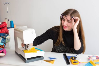 Teknoloji, ekipman, dikiş konsepti. Parlak kırmızı dudaklı genç bir kadın terzinin iş yerinde dikiş makinesiyle çalışıyor.