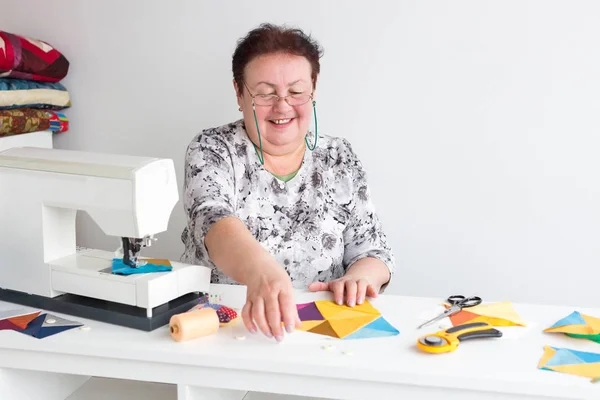 Trabajo, fabricación, concepto de acolchado. hay una costurera vieja y profesional en su mesa de trabajo con todas las herramientas que necesita y máquina de coser, ella está sosteniendo parches brillantes y sonriendo — Foto de Stock