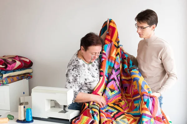 Quilting, artesanato, conceito de equipamento. no estúdio de costura há um homem jovem e uma mulher velha, eles estão segurando cobertor colorido feito por mãos na tecnologia original chamada patchwork — Fotografia de Stock