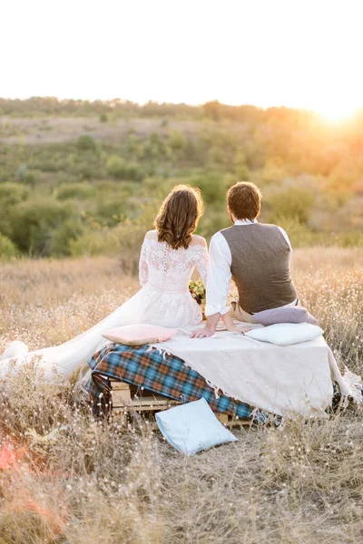 Visão traseira do jovem casal sentado em um campo e assistindo o pôr do sol  . — Fotografia de Stock