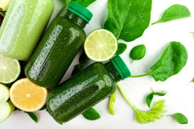 Taze bitki kökleri, elma, limon ve limon gibi çeşitli meyve ve sebzelerin arasında beyaz bir zemin üzerinde çeşitli yeşil içeceklerle dolu birkaç şişenin yaratıcı bir bileşimi..