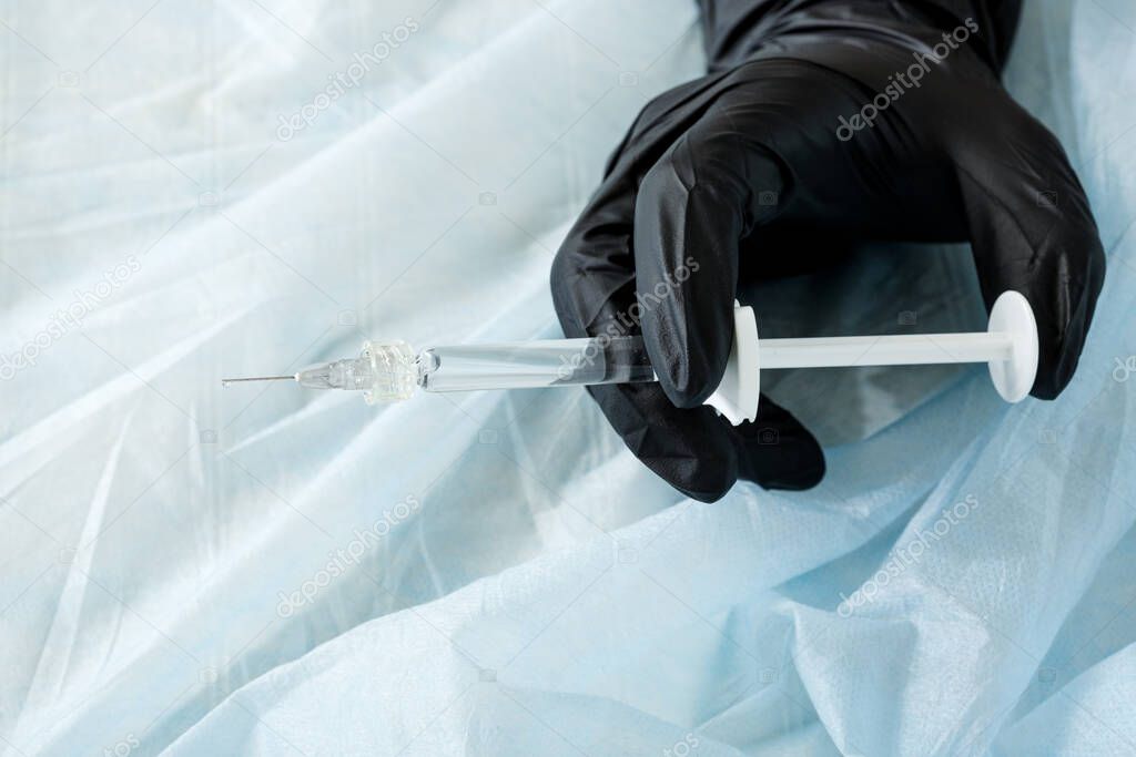 Close-up of the hand in a black latex glove with a syringe with a Luer lock on a blue spunbond fabric, an injection in a hospital. The vaccine against the virus.
