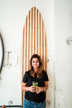 Kafede kahve içen hayalperest kadın. Çağdaş kafede elinde kahve fincanı olan rahat bir kadın tahta sörf tahtasının karşısında duruyor.