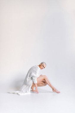 Beyaz örgü pelerinli zayıf kısa saçlı kadın kameraya bakıyor ve beyaz arka planda çömelmiş bacağını esnetiyor.