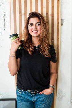 Kahveli neşeli güzel kadın. Bir fincan kahveyle kameraya gülümseyen, sıradan giyinmiş, sevimli mutlu bir kadın.