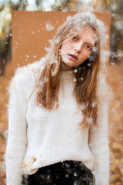 Güz ormanı arka planında başı eğik, kazaklı genç bir kadın etrafta uçuşan beyaz tüylerle