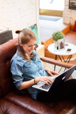 Kahvede dizüstü bilgisayarla takılan gülümseyen bir kadın. Rahat koltukta oturan rahat bir kadın ve mutlu bir şekilde dizüstü bilgisayar kullanıyor.