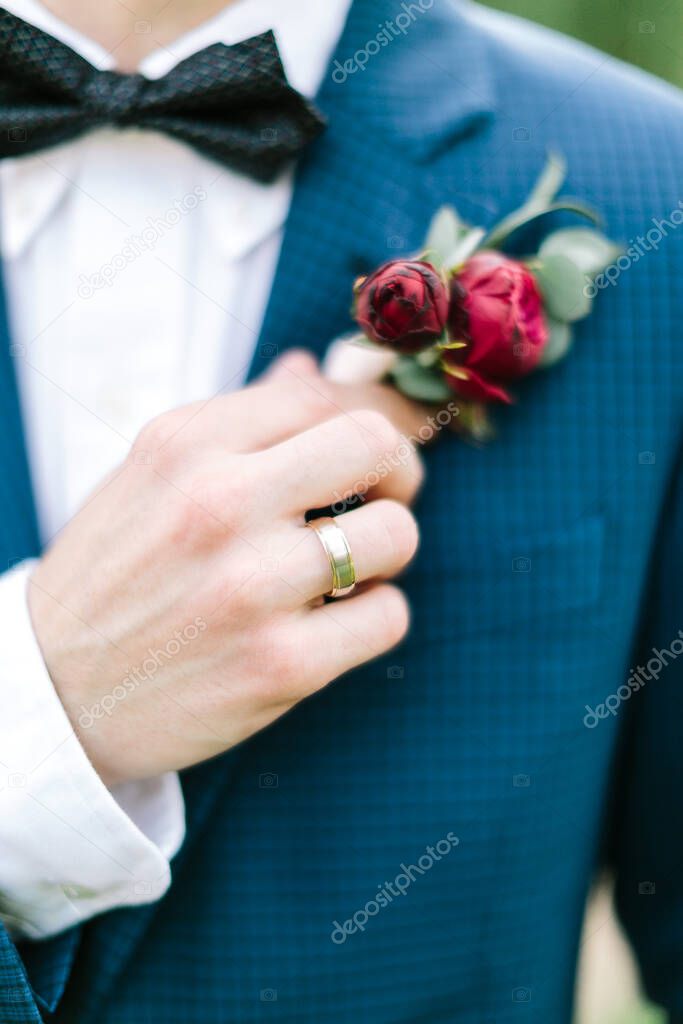 Soft focus of cropped groom's hand in wedding blue tuxedo and bowtie gently touches a boutonniere made of red peonies and eucalyptus