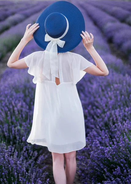 Una joven vestida con un vestido blanco y sosteniendo sus manos a un gran sombrero está caminando en medio de florecientes campos de lavanda. Marco vertical — Foto de Stock