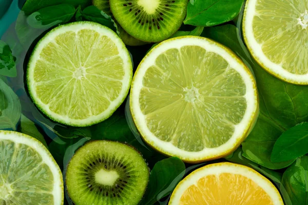 Grote platte ronde schijfjes verschillende citrusvruchten - limoen en citroen drijven samen met kiwi en groene bladeren in water. Het concept van tropische frisheid — Stockfoto