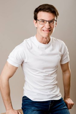 Genç, kaslı, gözlüklü, tişörtlü ve kot pantolonlu bir adam gri arka planda oturur ve kameraya bakarak kendisiyle dalga geçer..