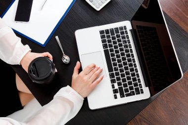 Masaüstünde yukarıdan bir manzara ve elinde bir fincan kahveyle dizüstü bilgisayarın üzerinde uzanan kadın elleri. İnternet araması, ağ iletişimi kavramı. Verilerle uzak çalışma