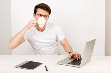 Genç profesyonel bir mühendis ya da girişimci, beyaz bir arka planda masasında oturmuş dizüstü bilgisayarla çalışırken çay içiyor. Çoklu görev kavramı ve dinlenme ve mola süresince uzaktan çalışma