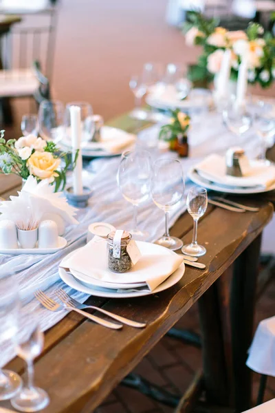 A line of served and decorated with dishes, glasses and flower spots at a festive wooden table. Side view. The concept of festive decorations.