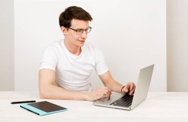 Tişört ve gözlük takan hafif gülümsemeli yakışıklı bir adam masasındaki beyaz bir duvara yaslanır ve bir dizüstü bilgisayarla etkileşime girer. Uzaktan karantina işi, eğitim ve iletişim kavramı