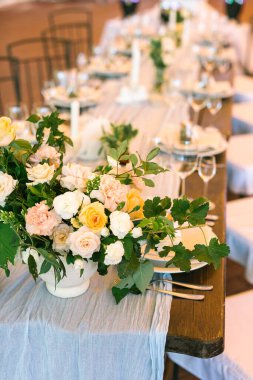 Dikey çerçeve, üst görünüm. Merkez çiçek aranjmanına odaklanıyoruz, doğal bir kumaşa yerleştiriliyor, festival masasını örtüyor, tabaklar ve bardaklarla güzelce servis ediliyor..