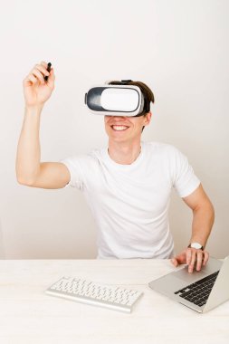 Sanal gerçeklik kulaklığı kullanan genç bir laboratuvar asistanı, beyaz arka planda klavye ve dizüstü bilgisayarla birlikte masasında otururken üç boyutlu uzayda yönlendirilmiş nesnelerle etkileşime giriyor.. 