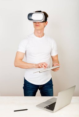 Sanal gerçeklik kulaklığı kullanan genç adam üç boyutlu uzayda yönlendirilmiş şeylerle etkileşime girerken masasında klavye ve dizüstü bilgisayarla beyaz arka planda oturuyor.. 
