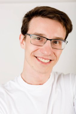 Siyah saçlı, gözlüklü ve tişörtlü, beyaz arkaplanda özçekim yapan genç gülen adam..