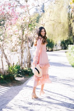 Yumuşak odaklanma. Uzun saçlı, zarif şapkalı, gülümseyen kadın güneşli bahçe yolunda yürürken omzunun üzerinden kameraya bakıyor.