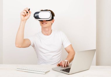 Genç adam Grafik Tasarımcı sanal gerçeklik kulaklığı kullanarak üç boyutlu uzayda hareket ederken, beyaz arkaplanda klavye ve dizüstü bilgisayarla birlikte oturuyor.. 