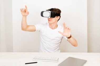 Sanal gerçeklik kulaklığı kullanan genç bir astrofizikçi, beyaz arka planda klavye ve dizüstü bilgisayarla birlikte masasında otururken üç boyutlu uzayda yönlendirilmiş şeylerle etkileşime girer.. 