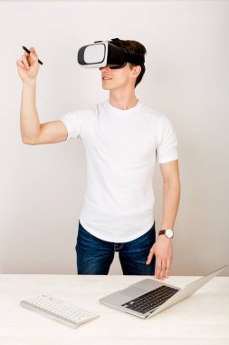 Sanal gerçeklik kulaklığı kullanan genç bir iç mimar, beyaz arka planda klavye ve dizüstü bilgisayarla birlikte masasında otururken üç boyutlu alanda yönlendirilen artırılmış nesnelerle etkileşime giriyor.. 