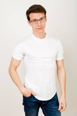Sıradan giyinmiş çekici bir adam beyaz bir arka planda duruyor, eli cebinde ve kameraya bakıyor, düşünceli, ama sakin ve özgüvenli bir şekilde..