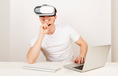 Sanal gerçeklik kulaklığı kullanan genç bir öğrenci, beyaz arka planda klavye ve dizüstü bilgisayarla birlikte masasında otururken üç boyutlu uzayda yönlendirilen artırılmış şeylerle etkileşim halindedir.. 