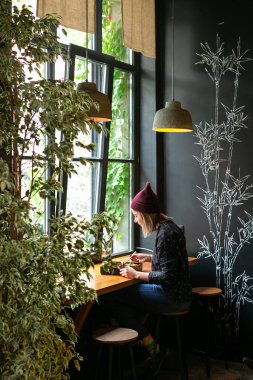 Gündelik kıyafetli genç bir kız büyük pencerenin arkasındaki ahşap bir tezgahın arkasında tek başına oturuyor ve düşünceli bir şekilde bir tabak yemeğe bakıyor. Dikey çerçeve, yan görünüm.