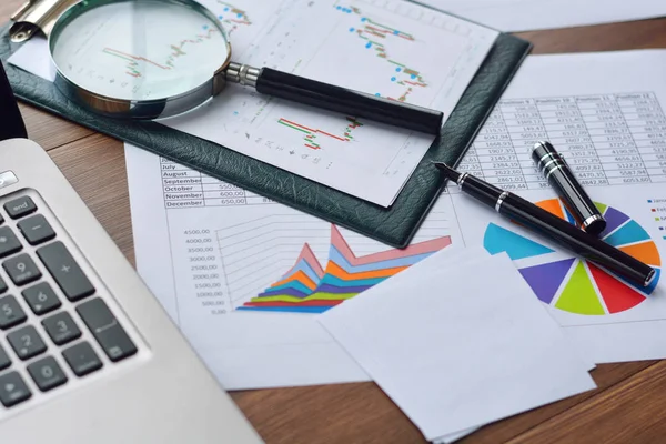 Financial printed paper charts, graphs, diagrams, magnifying glass, pen and computer on wooden table.