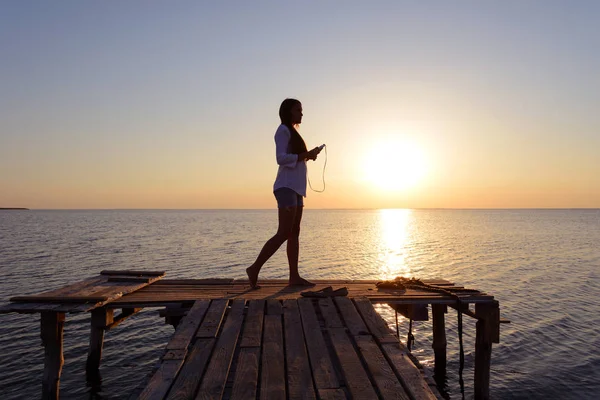 Feliz niña alegre en una camisa blanca escuchando música en los auriculares y bailando en el muelle junto al mar al atardecer — Foto de Stock