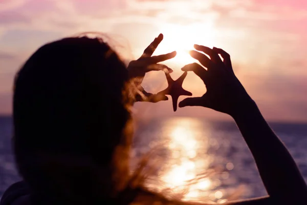 Joven, hermosa con el pelo largo volando la niña sostiene la estrella de mar en sus manos en el fondo de la puesta de sol en la orilla del mar. Romántico verano mar fondo — Foto de Stock