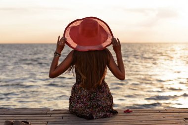 Beautiful young slender girl in sarafan and long hair and coral hat sits on pier (bridge) by sea (ocean) and looks at sunset (dawn) of  sun. Thoughtful and dreaming stranger on a summer evening clipart