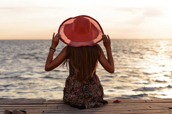 Beautiful young slender girl in sarafan and long hair and coral hat sits on pier (bridge) by sea (ocean) and looks at sunset (dawn) of  sun. Thoughtful and dreaming stranger on a summer evening