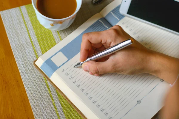 Bloco de notas, caneta, laptop (computador), telefone e uma xícara de café na mesa. caligrafia, mão escreve uma caneta em um caderno . — Fotografia de Stock