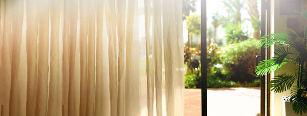 Тропический фон. Лето, путешествия, отпуск и отдых. Открытое окно, дверь и белый занавес с размытым видом на пальмовый сад из отеля. Экзотические растения в солнечный день, скопируйте пространство. Баннер
