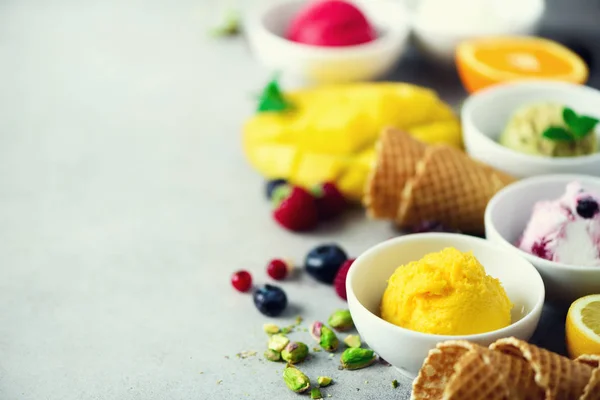 Ice cream balls in bowls, waffle cones, berries, orange, mango, pistachio on grey concrete background. Colorful collection, flat lay, summer concept — Stock Photo, Image