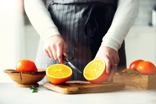 Manos de mujer rebanando naranja, cortando cítricos. Cuchillo, tabla de cortar de madera en la cocina blanca de diseño. Copiar espacio — Foto de Stock