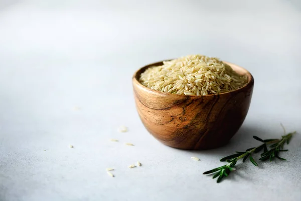 Rauwe biologische bruine rijst in houten kom en rozemarijn op lichte concrete achtergrond. Voedingsmiddelen en nieuwe voedselingrediënten. Kopiëren van ruimte — Stockfoto