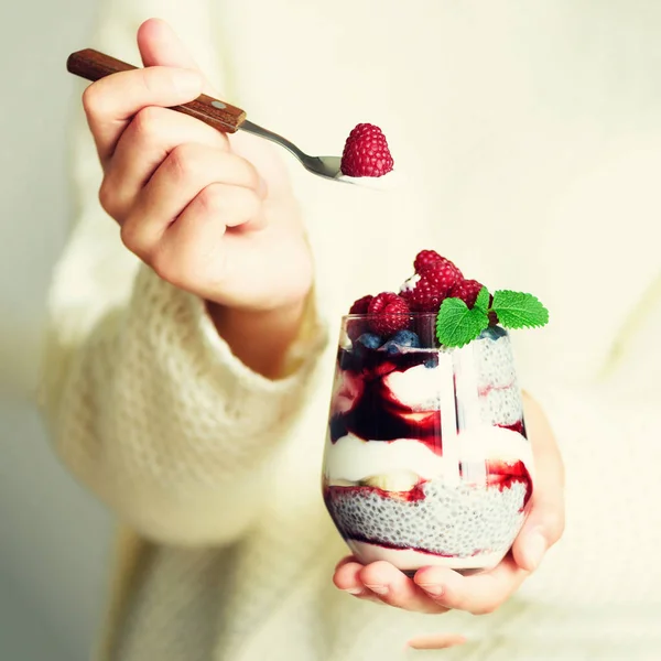 Chica en suéter blanco sostiene vidrio con postre de leche de chía, frambuesas, arándanos, menta. Concepto de desayuno saludable. Copiar espacio, cerrar. Mañana soleada. Cultivo cuadrado — Foto de Stock