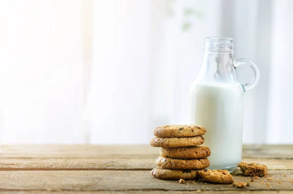 Chocolate chip cookies, bottle and glass of milk on wooden table near window, white background. Sunny morning, copy space — Stock Photo, Image