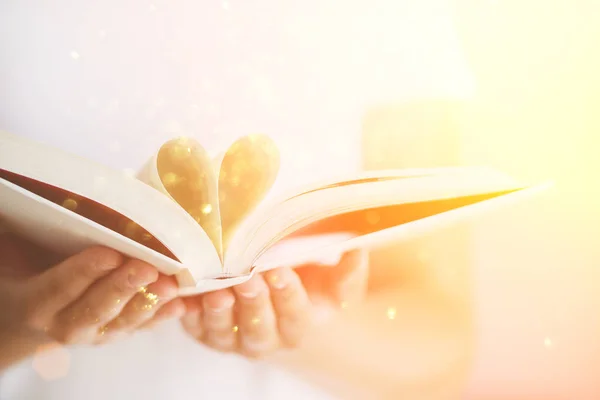 Livro com páginas abertas e forma de coração em mãos de menina. Espaço para cópia. Conceito de amor. Fundo festivo com bokeh e luz solar. Conto de fadas mágico — Fotografia de Stock